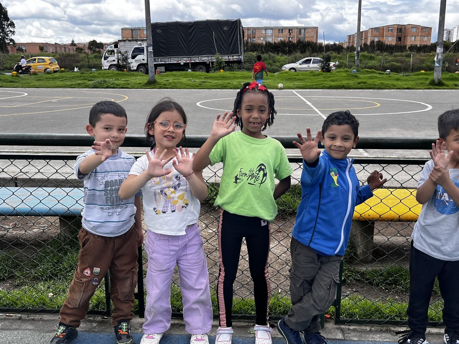 Un grupo de niños y niñas sonríe y saluda con la mano junto a una cancha al aire libre; una de las niñas lleva la camiseta verde de Formarte.