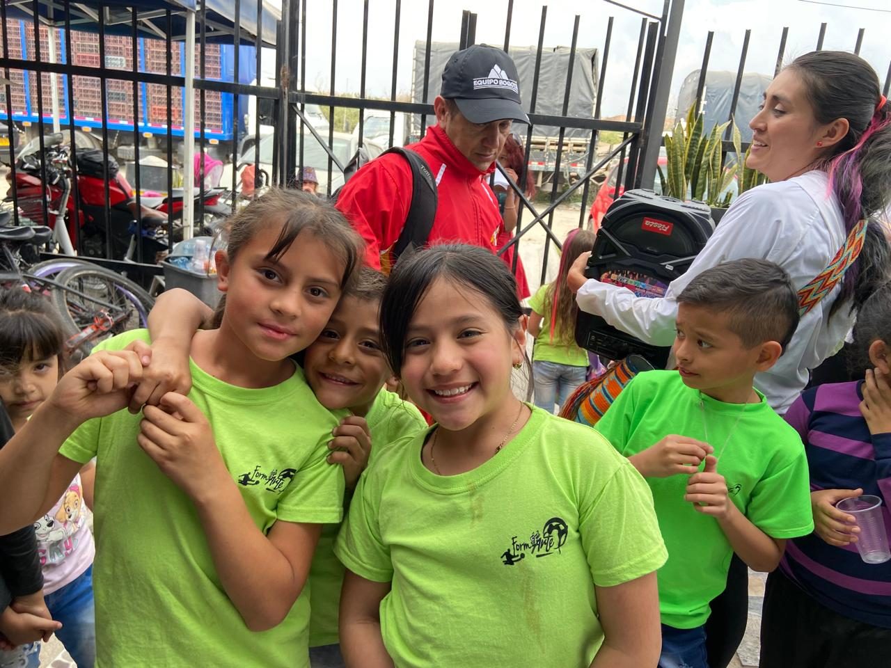 Un grupo de niños y niñas con camisetas verdes de la fundación sonríe y se abraza durante una jornada al aire libre, con voluntarios detrás.