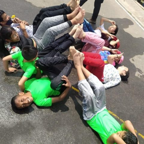 Niños y niñas con camisetas verdes juegan tendidos en el suelo durante una actividad deportiva al aire libre.